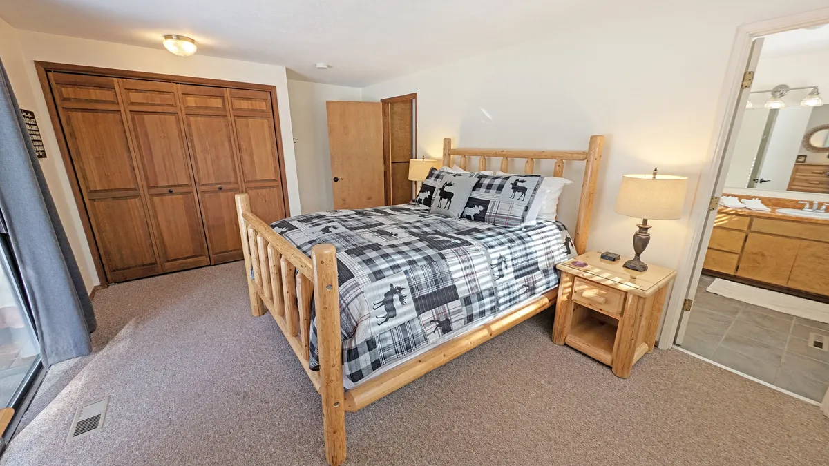 A serene bedroom featuring a rustic log bed with plush plaid bedding, a nightstand with a softly glowing lamp, and closet doors. Natural light pours in from a nearby window, creating a peaceful retreat ideal for relaxing and unwinding.