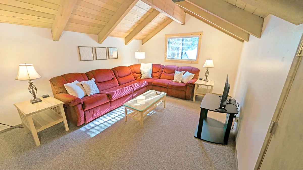 A warm, spacious loft area featuring a large red sectional sofa arranged for conversation or lounging, a glass coffee table at the center, a TV stand with flatscreen TV opposite the sofa, and wooden beams accenting the ceiling. A cozy reading nook can be glimpsed near the window, with a bookshelf stocked with books, enhancing the inviting, multipurpose feel of the space.