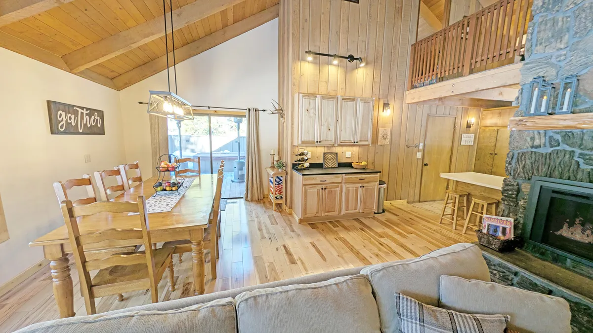 This interior image captures the spacious dining table set near the kitchen, with a fireplace and sliding glass door visible. The room is filled with natural light, and the layout showcases the open flow between the living, dining, and kitchen areas. Comfortable seating and the warmth from the fire create an inviting atmosphere for family gatherings.