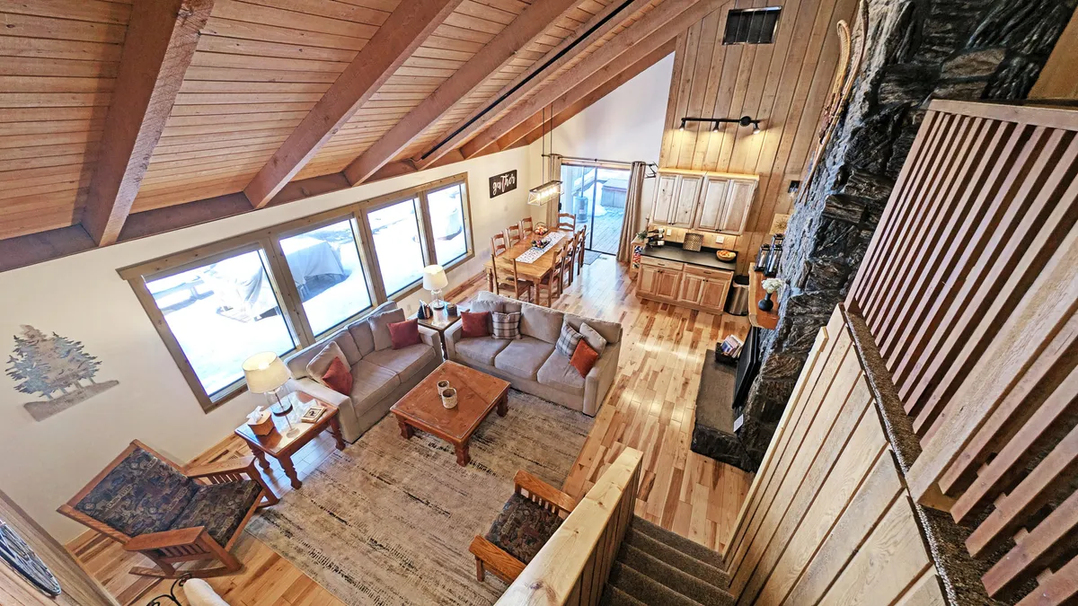 A spacious, light-filled common area boasting large windows with abundant natural light, a comfortable seating area, a wooden dining table, and a prominent stone fireplace. The view through the windows hints at a connection to the outdoor environment, enhancing the indoor-outdoor living experience.