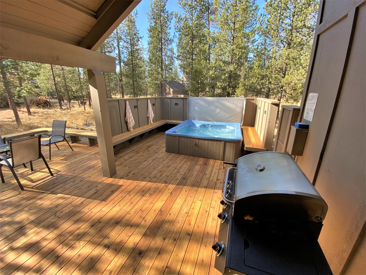 An exterior view of the back deck, showing the hot tub, gas grill, outdoor seating, and towels, surrounded by nature. The sliding doors leading inside suggest an easy indoor-outdoor flow, perfect for barbecues and relaxing after family meals.
