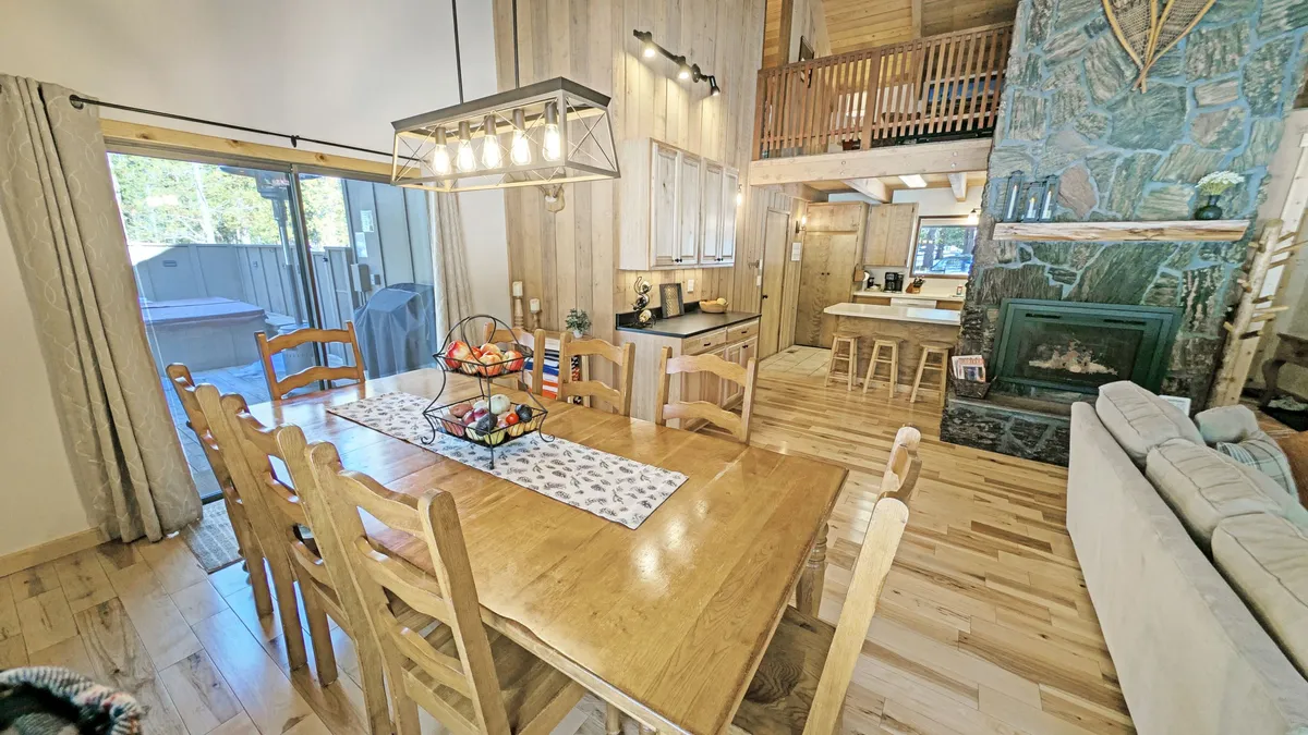 A warm, inviting dining area featuring a wooden dining table, a cozy fireplace, a view into the kitchen, and large windows that hint at outdoor light and scenery. The space is set for gatherings and meals, with comfortable seating and an open flow to the kitchen.