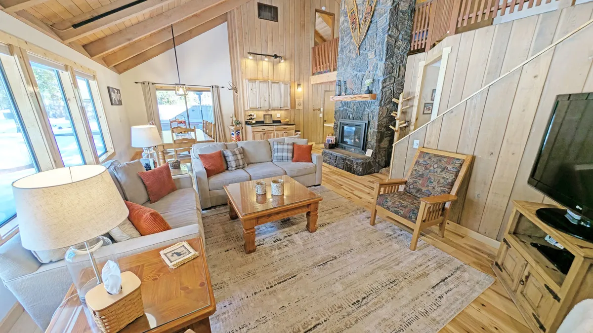 The image captures a spacious living area with a striking stone fireplace, exposed wooden beams, a comfortable seating arrangement, large windows flooding the room with natural light, and a decorative rug. The space is ideal for relaxing, reading, or socializing, embodying the cozy, modern ambiance described in the section.