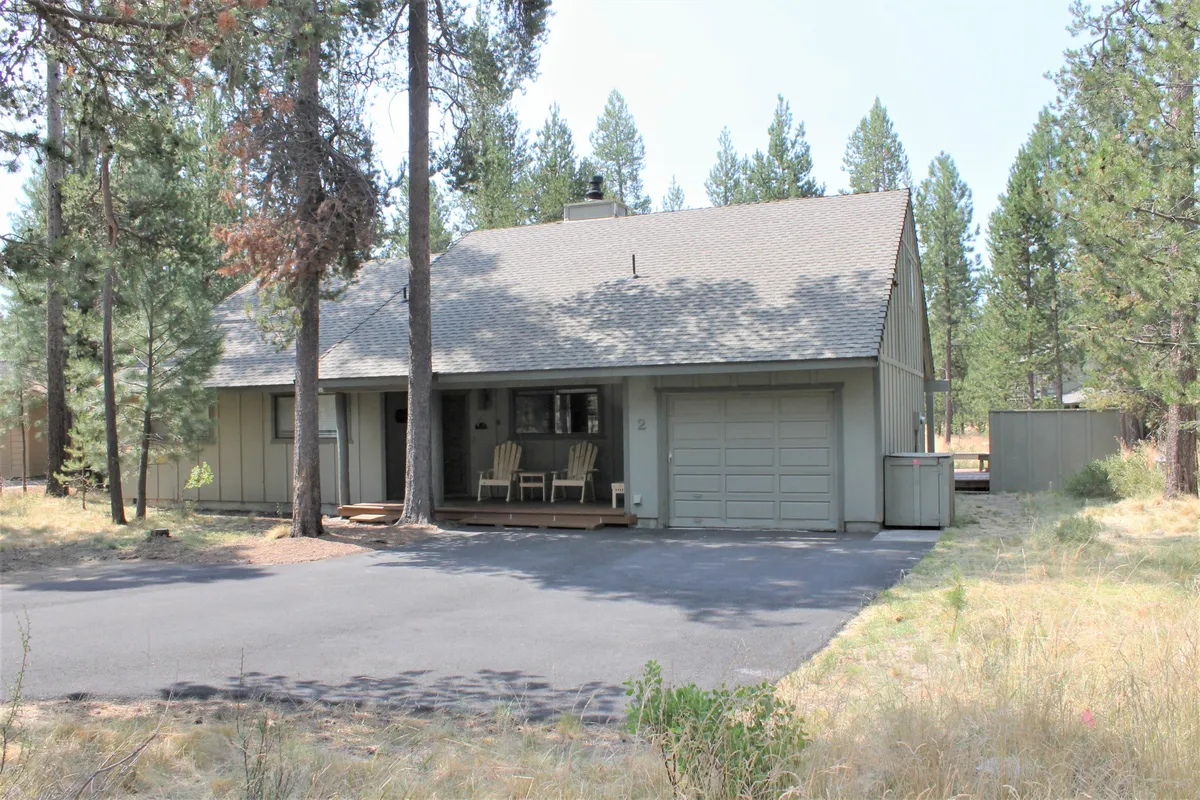 A tranquil exterior view of the home's front porch framed by trees, with the driveway and garage visible. The scene suggests a peaceful setting perfect for relaxing, gathering, or launching outdoor adventures.