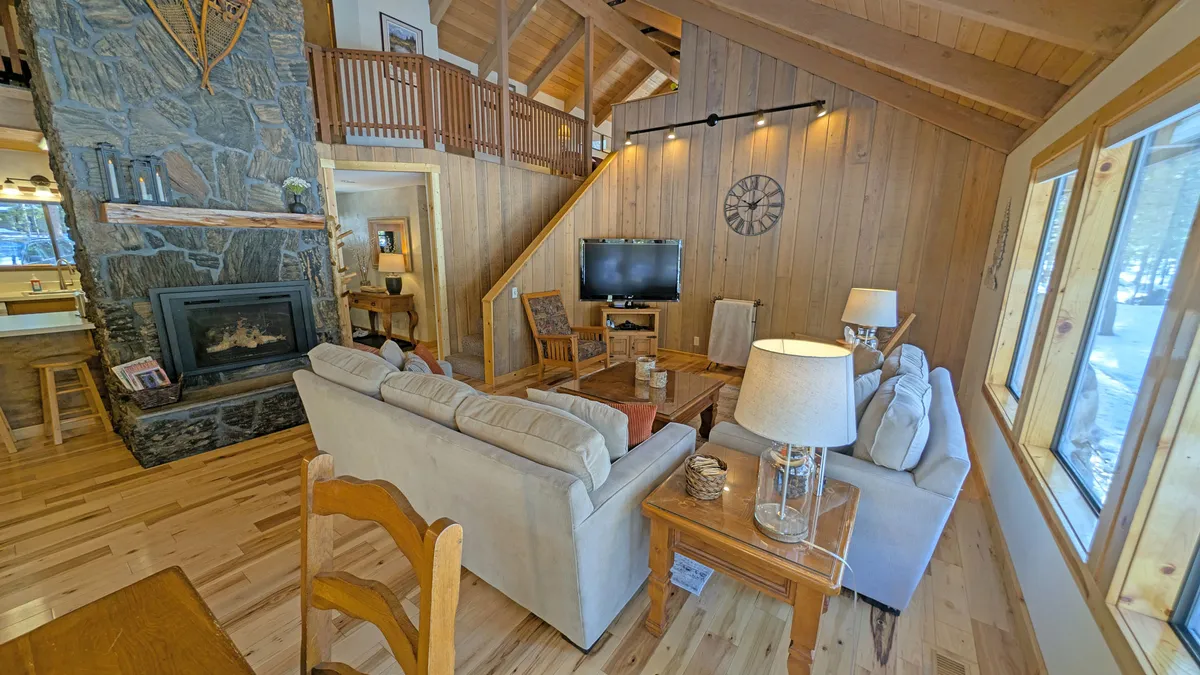 A warm and spacious living area with a fireplace, sofa seating, wooden accents, and large windows letting in abundant natural light. The setting invites relaxation and family connection.