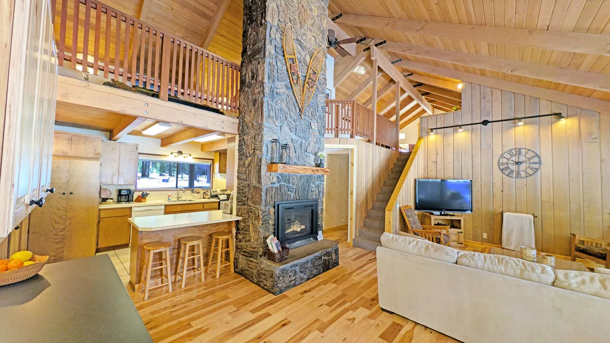 The hero image showcases a spacious, warmly lit living area with a striking stone fireplace, rustic wooden beams, comfortable sofas, and an open kitchen layout, inviting guests to gather and relax. A sleek balcony railing overlooks the space, emphasizing both coziness and openness—perfect for family activities and group connection.